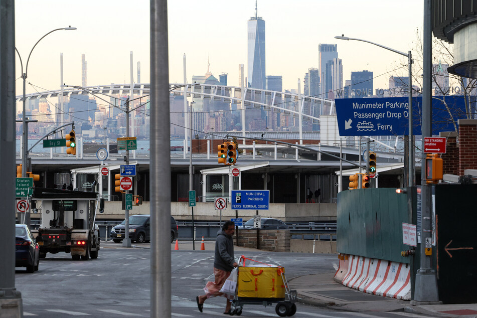 A man crosses a street in the Tompkinsville neighborhood of Staten Island, with the Manhattan skyline visible in the distance, on January 23, 2026.