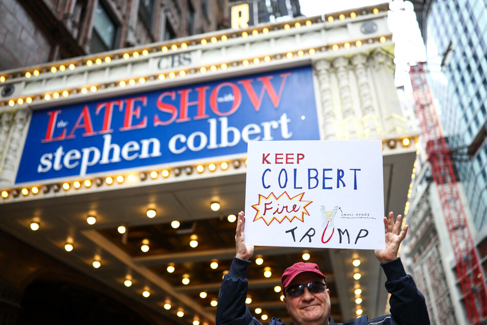 A man holds a "Keep Colbert, Fire Trump" sign outside the Ed Sullivan Theater ahead of the taping of the final episode of the The Late Show on May 21, 2026.