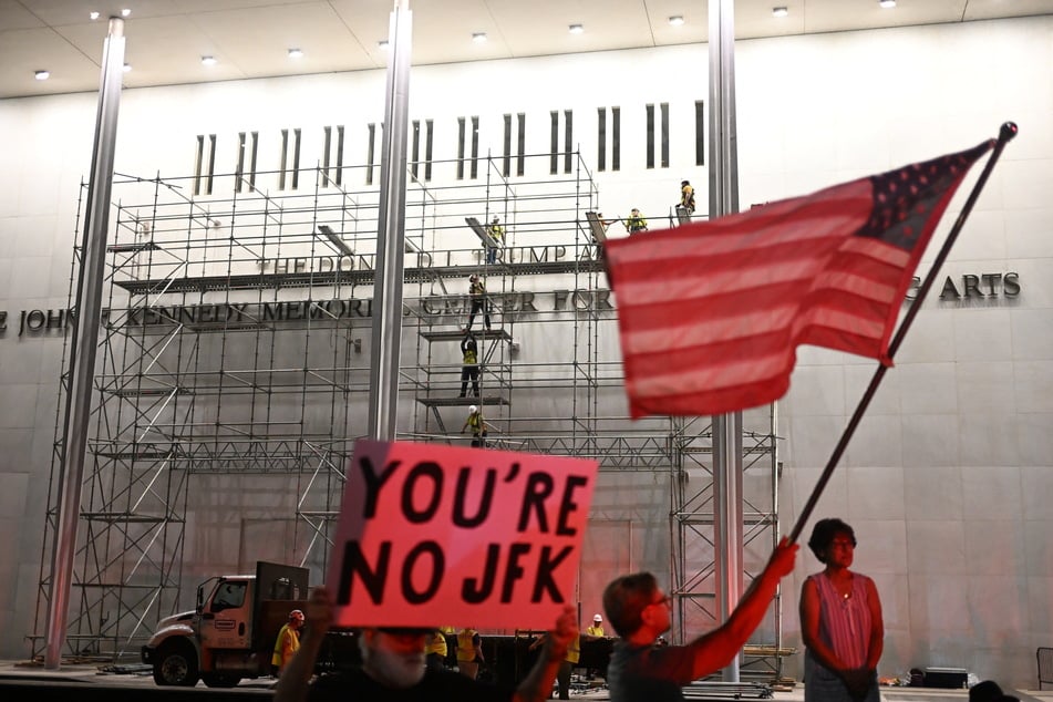 Protesters wave a US flag and hold a sign reading "You're no JFK" as workers build scaffolding near the signage for the "Donald J. Trump and John F. Kennedy Memorial Center for the Performing Arts" on June 12, 2026.