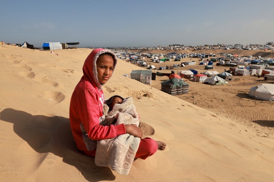 A Palestinian girl sits holding a toddler on a sand dune overlooking a camp for displaced people in Rafah in the southern Gaza Strip.