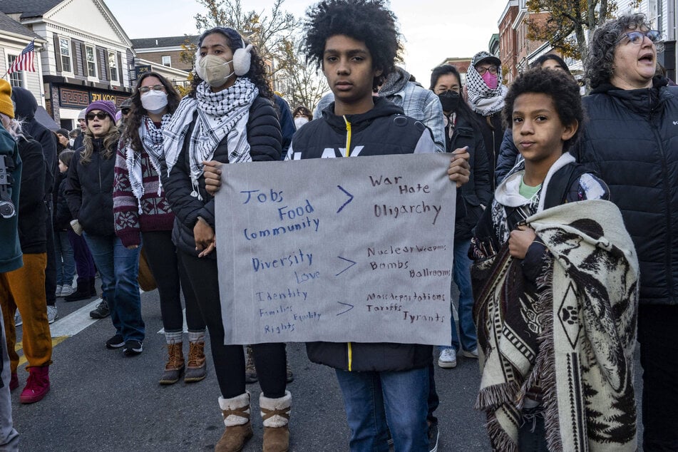 Youth hold a sign calling for "jobs," "food," and "community" over "war," "hate," and "oligarchy" while other participants wear keffiyehs during the 2025 National Day of Mourning.