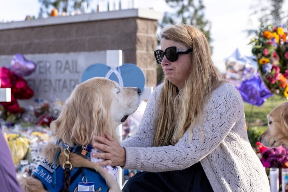 A woman pets a therapy dog at a memorial site outside of Silver Rail Elementary School in Bend, Oregon, on October 17, 2026. A suspected16-year-old shooter killed three children and one teacher, injuring others, at the school on Thursday, and parents are figuring out. how to talk to their kids about what happened. 