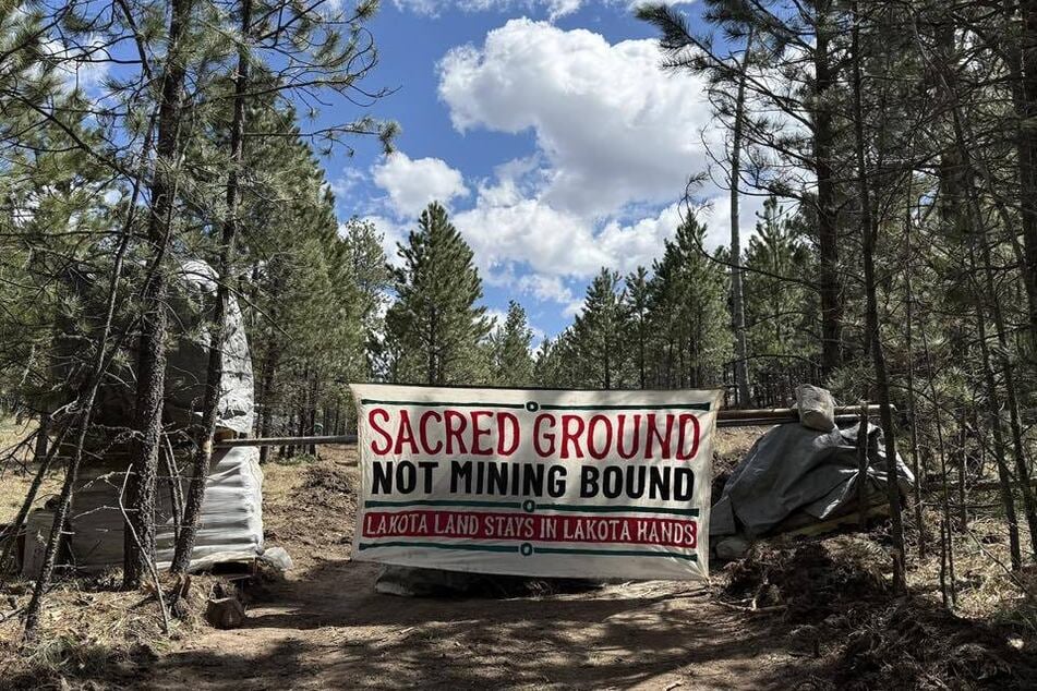 A banner reading "Sacred Ground Not Mining Bound" is pictured as Indigenous groups resist graphite drilling in the Black Hills.