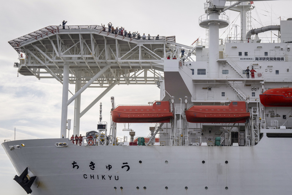Crew members wave to the watching public from aboard Japan's deep-sea drilling vessel Chikyu as it returns to Shimizu port on February 14, 2026.
