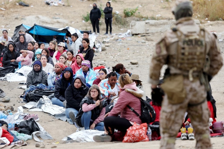 Immigrants seeking asylum in the US, who are stuck in a makeshift camp between border walls between the US and Mexico, sit as a Customs and Border Protection officer keeps watch.
