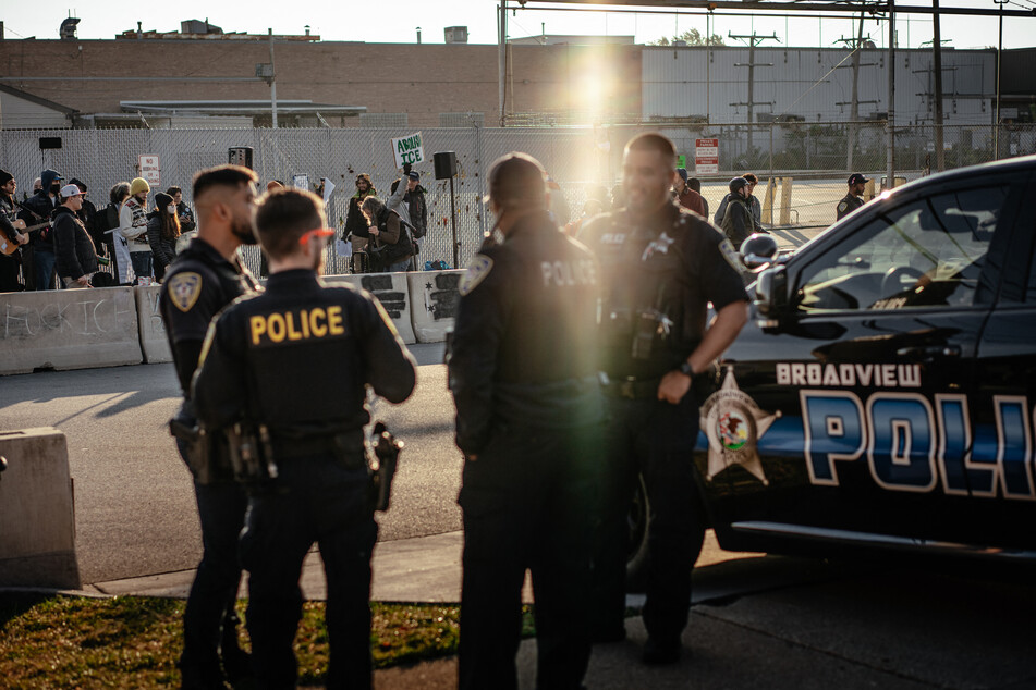 Local police officers watch from across the street as demonstrators gather and sing in the "free speech zone" outside the ICE processing and detention facility in Broadview, Illinois, on October 31, 2025.