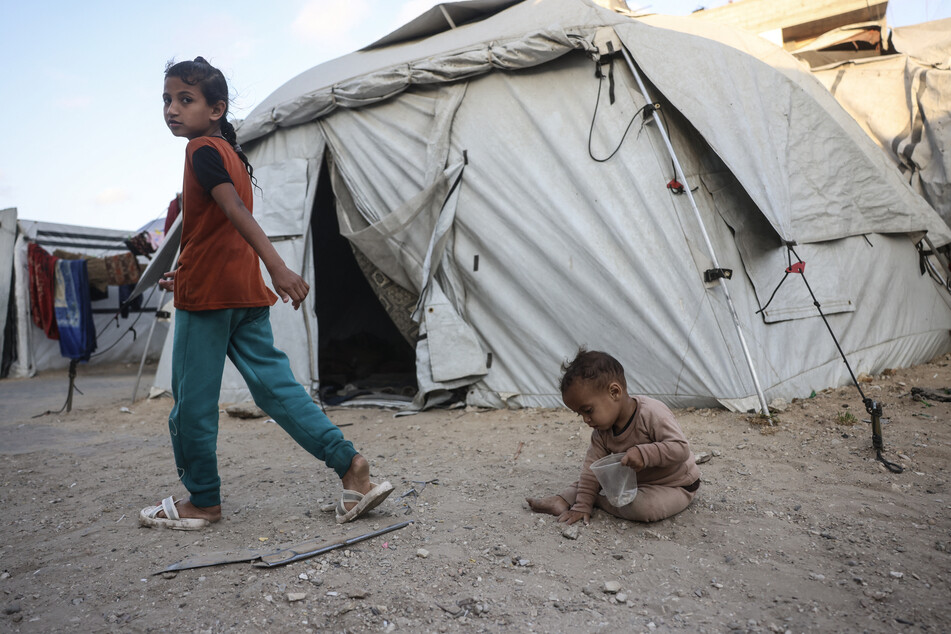 A Palestinian girl walks past a toddler playing among makeshift shelters in Khan Younis in the southern Gaza Strip on May 21, 2026.